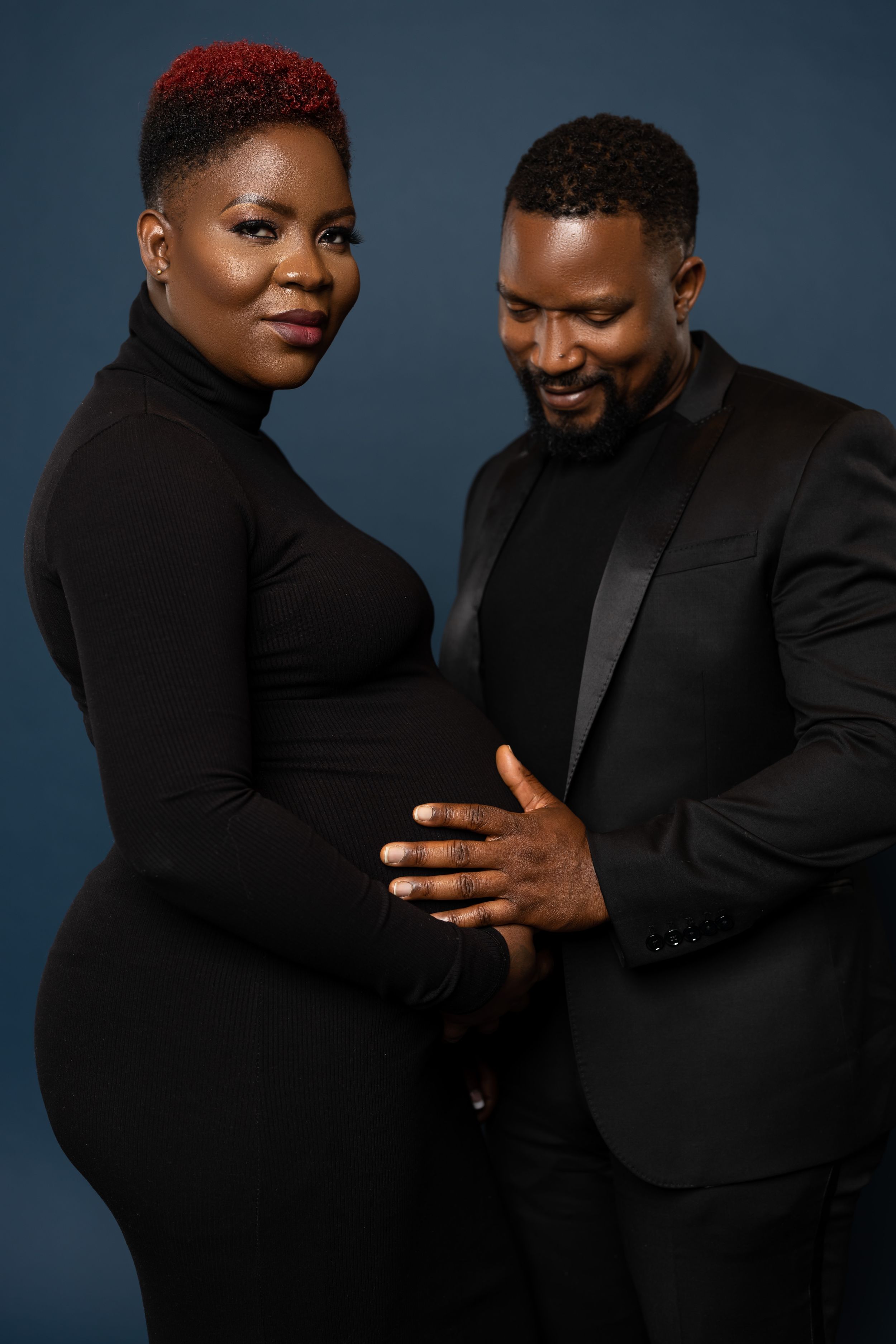 A couple poses against a dark background both wearing black for an elegant maternity photo.