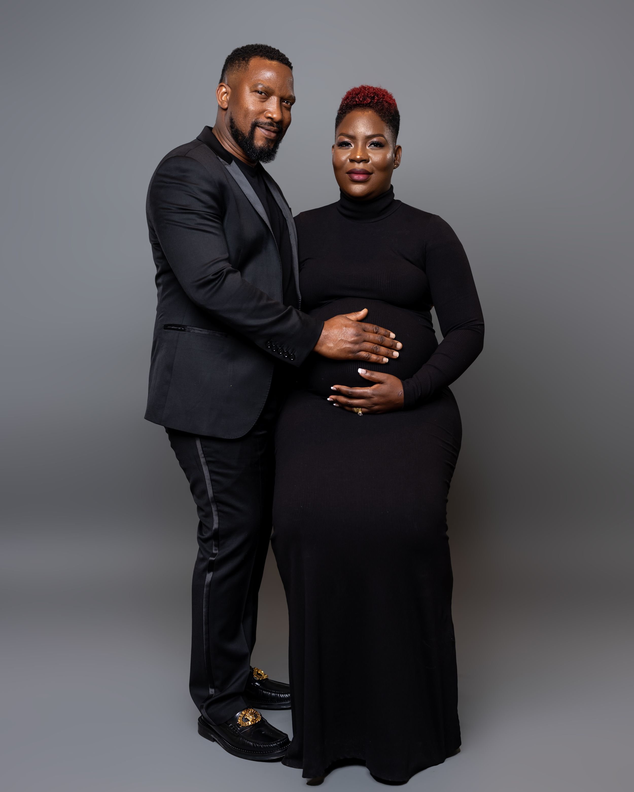 Elegant studio portrait of couple in formal black attire against grey backdrop.