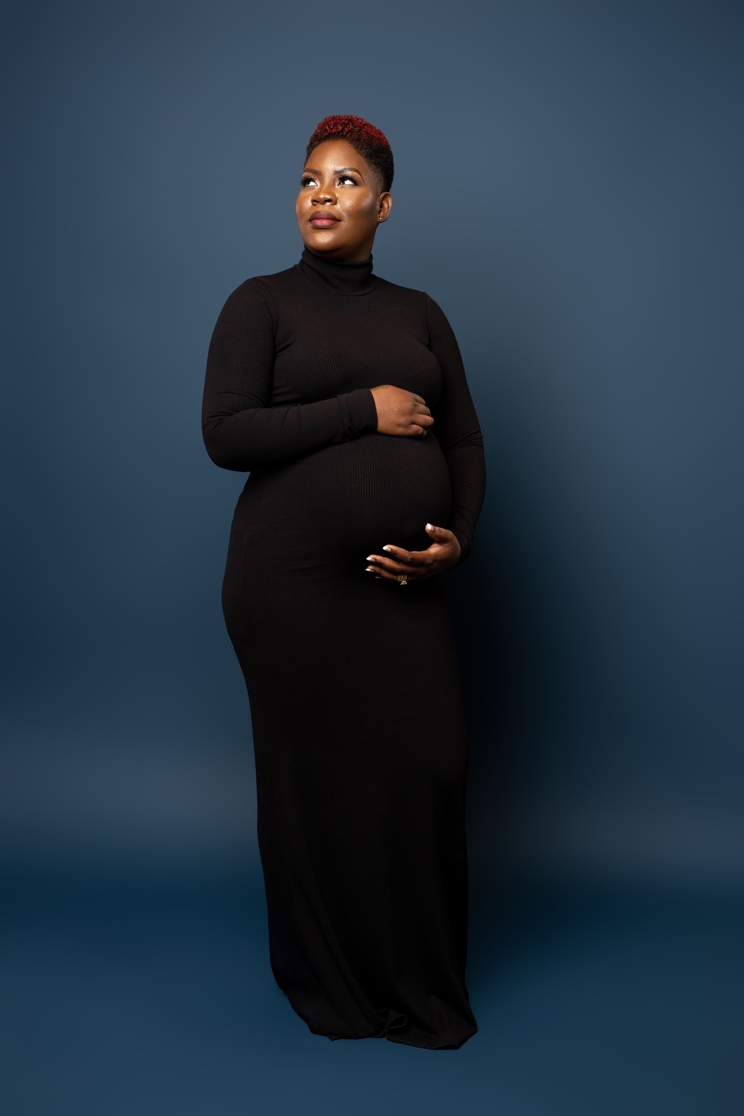 Full-length maternity portrait of woman in black dress against dark blue background.