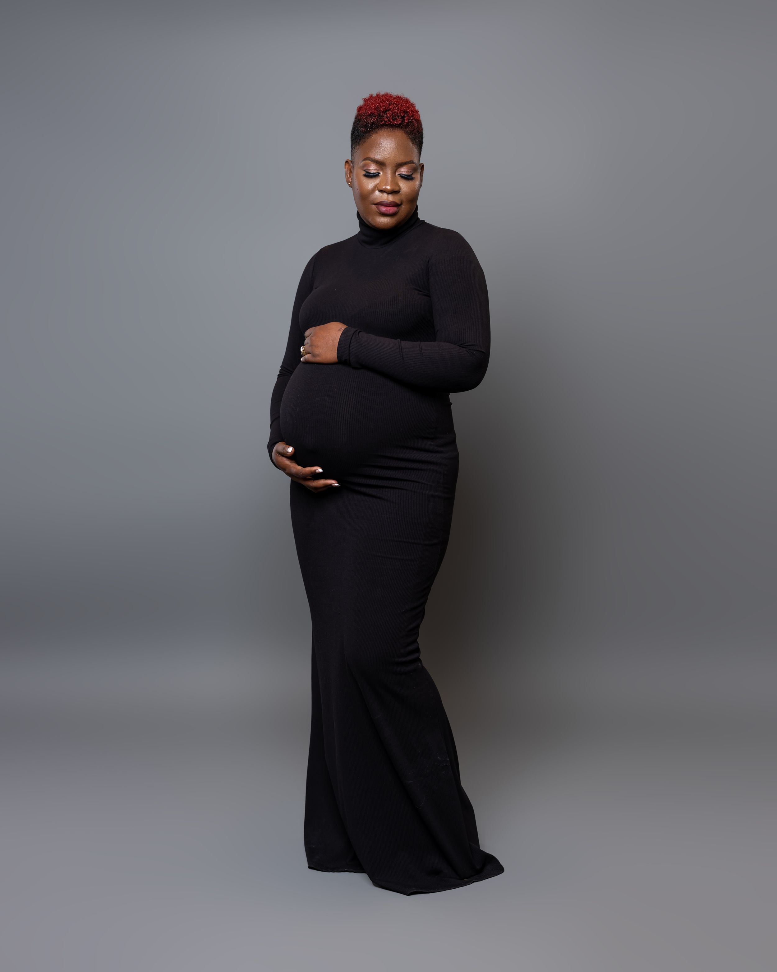 Maternity portrait of woman in black dress posing against grey background.
