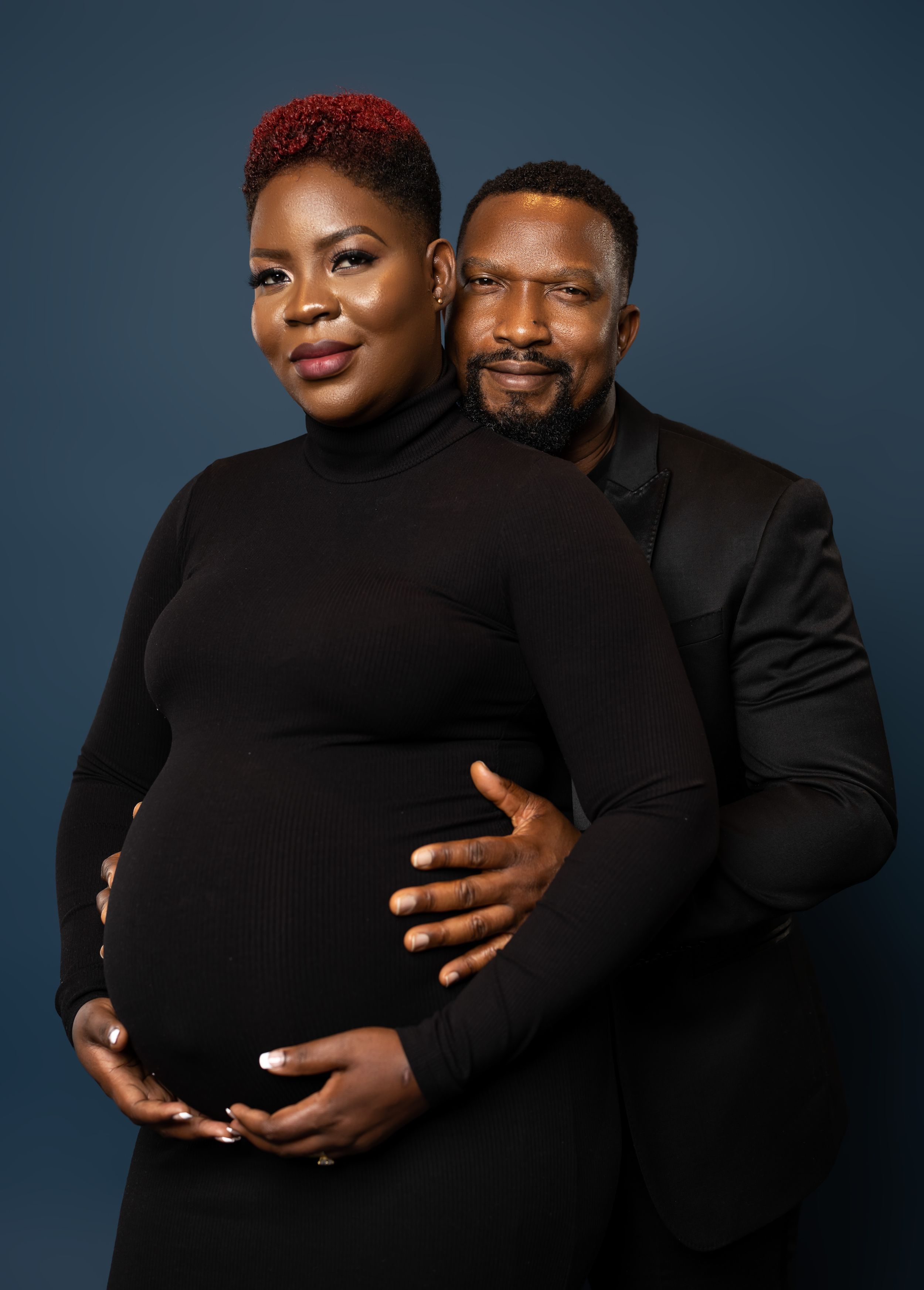 Intimate maternity portrait of couple in black clothing against dark backdrop.