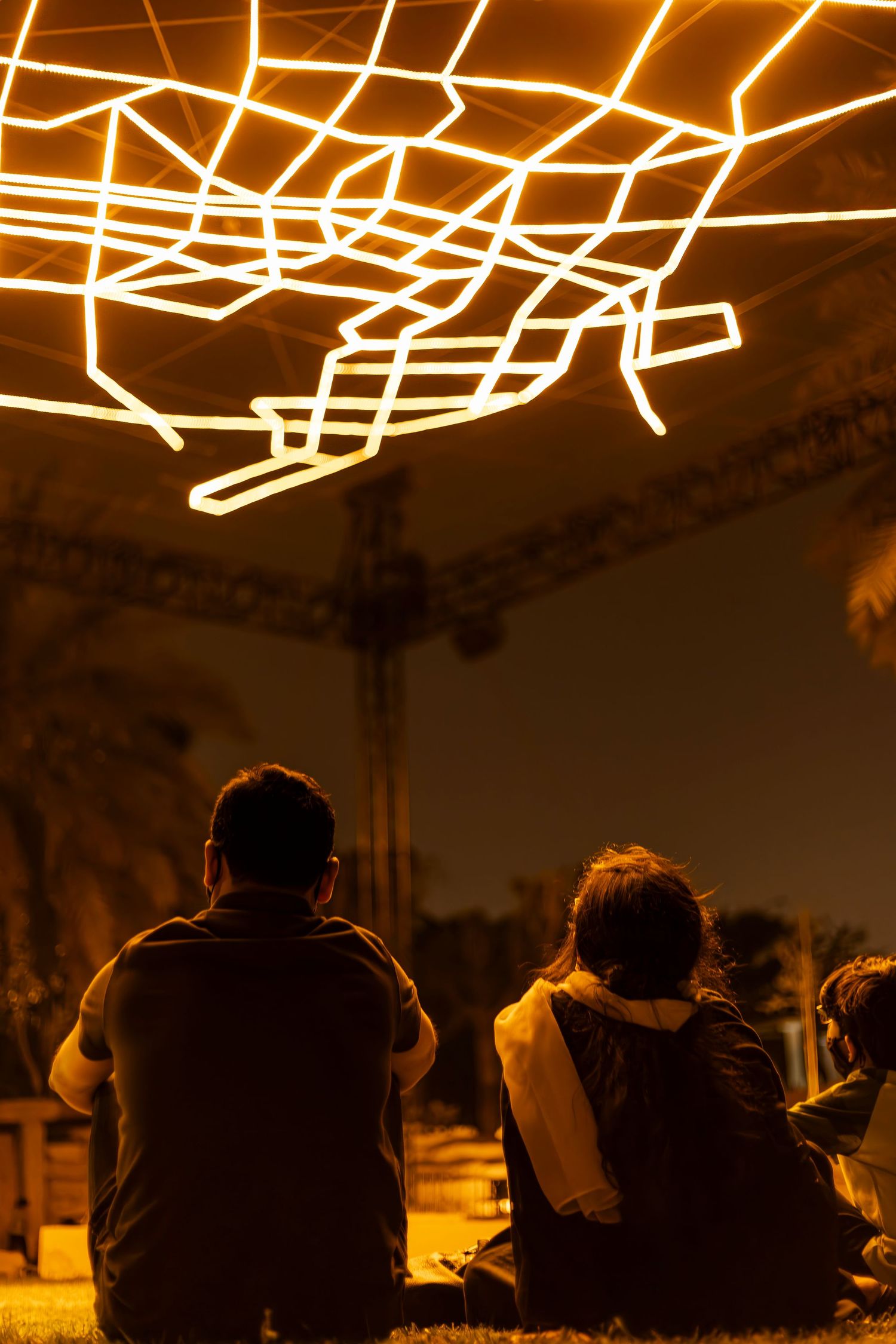 Silhouettes of people sit beneath glowing geometric light patterns in a warm nighttime atmosphere.