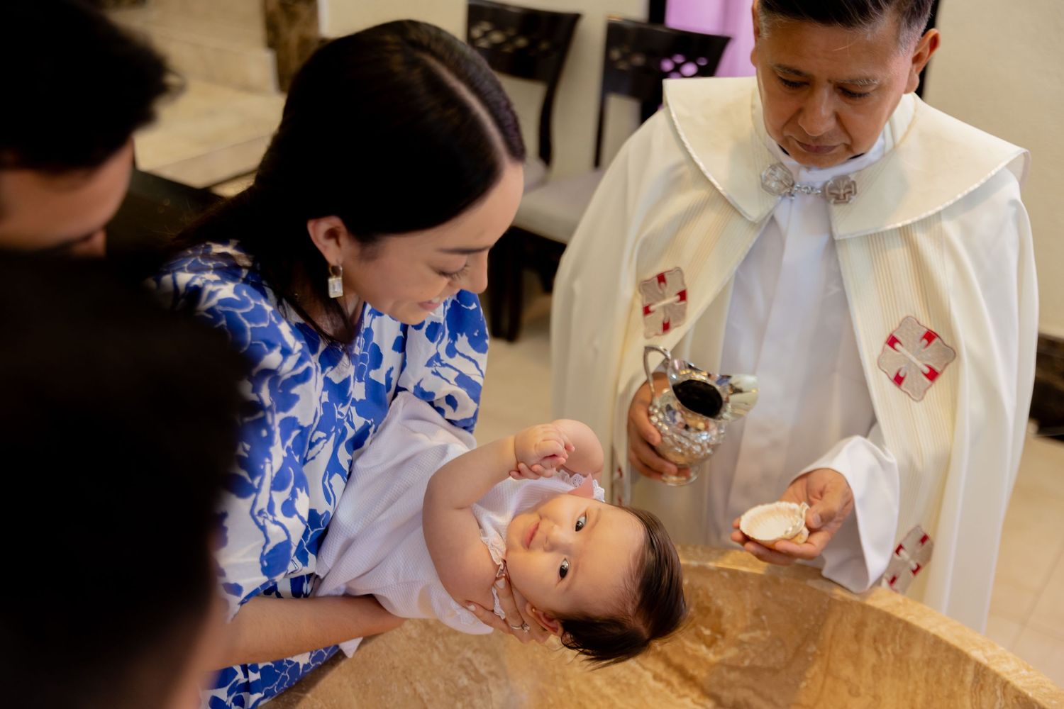 Catholic priest baptizes a baby at a baptismal font while mother holds the infant.