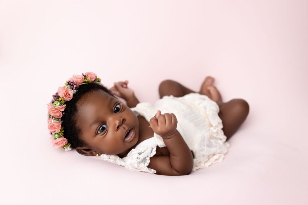 Baby wearing white dress and floral crown lying on pink background.