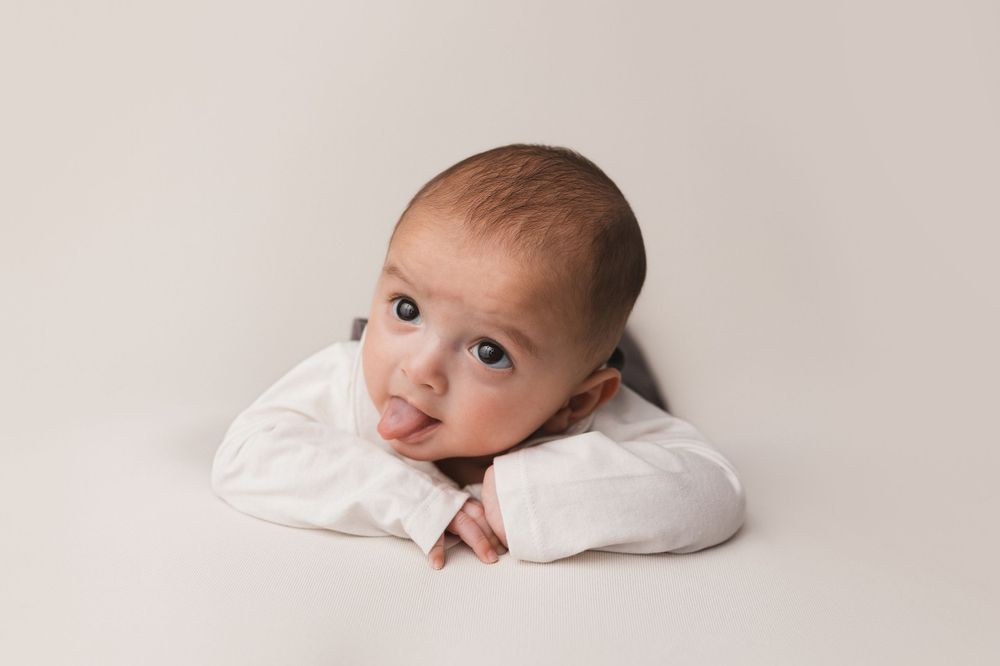 Baby in white outfit resting head on hands against neutral background.