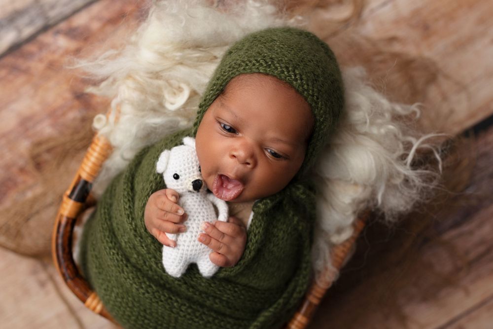 Smiling baby wearing green knit hat lying in basket with white plush toy.