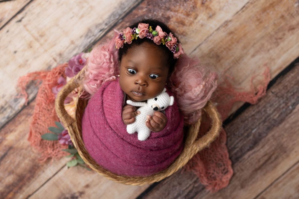 Newborn wrapped in purple swaddle with floral crown lying in wooden basket.