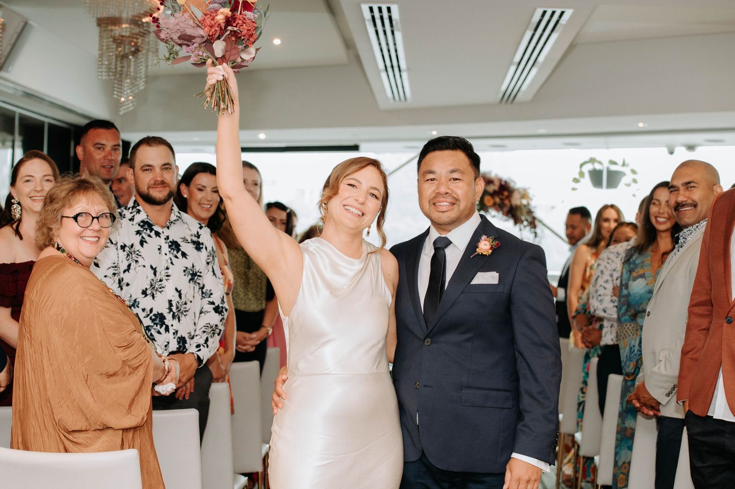 A couple celebrates their wedding day surrounded by guests as they raise a celebratory toast indoors.