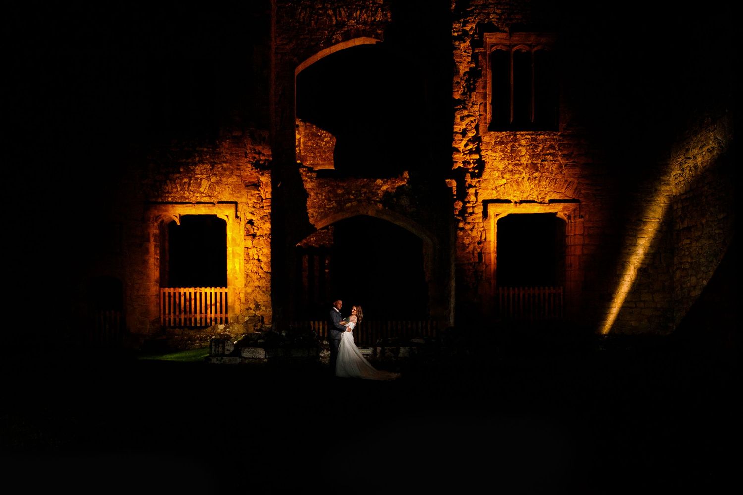Dramatic nighttime wedding photo with couple in archway of illuminated stone building.