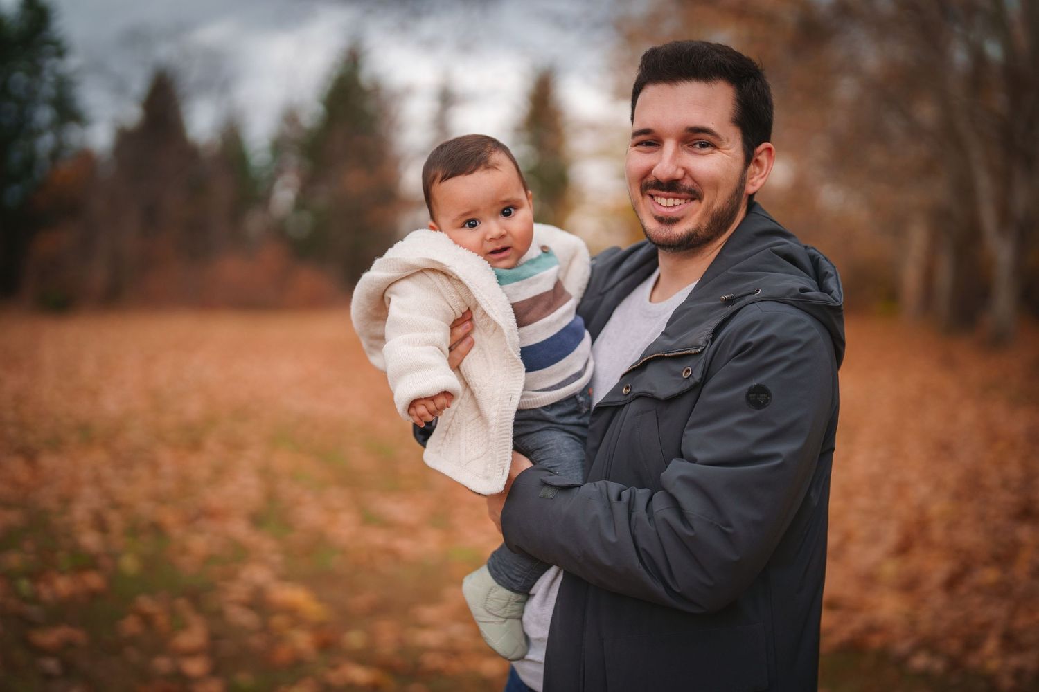 Parent holding smiling baby outdoors during autumn photo session.