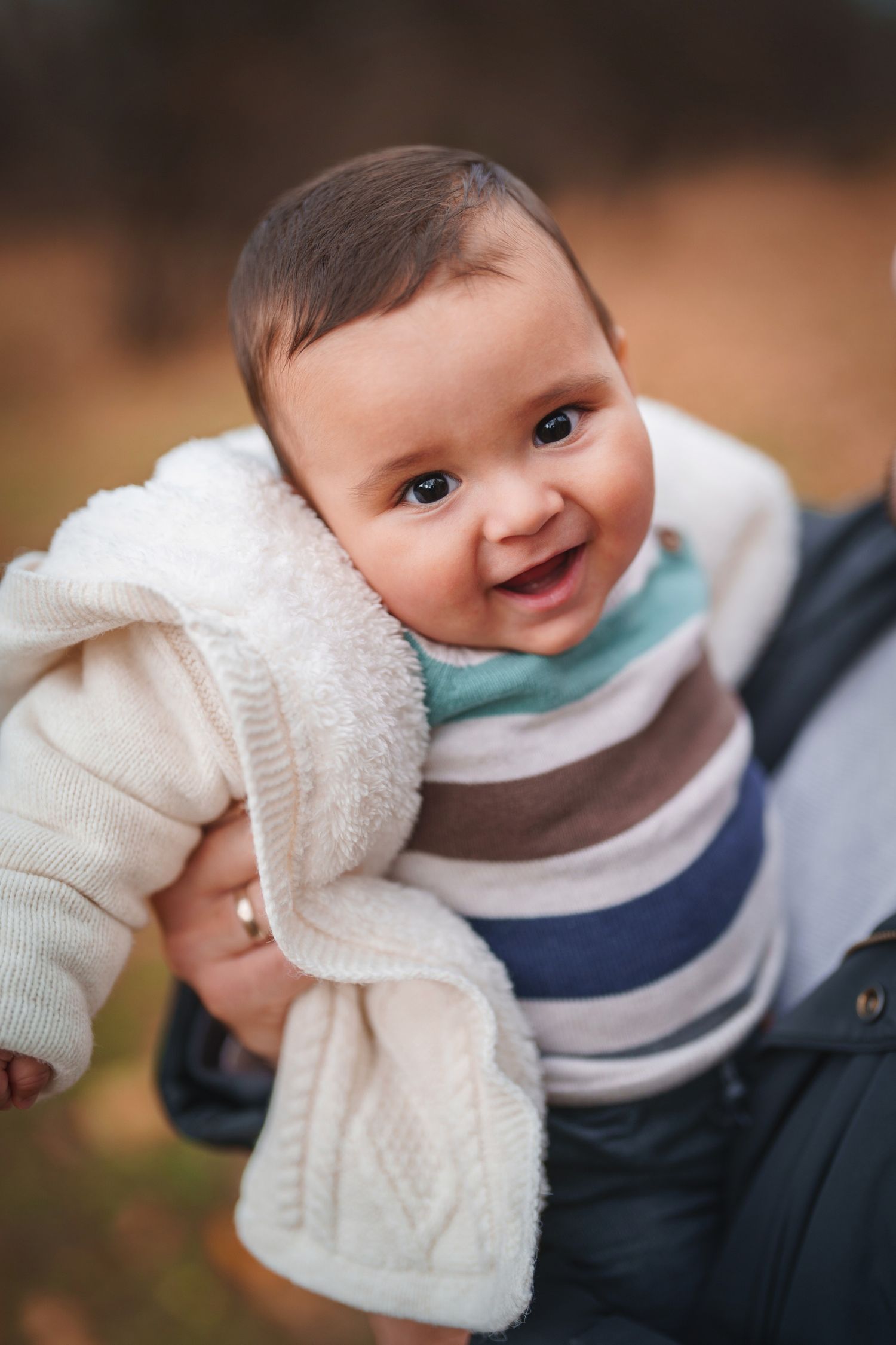 Baby wearing striped sweater and white cardigan smiles brightly at camera during autumn photoshoot.