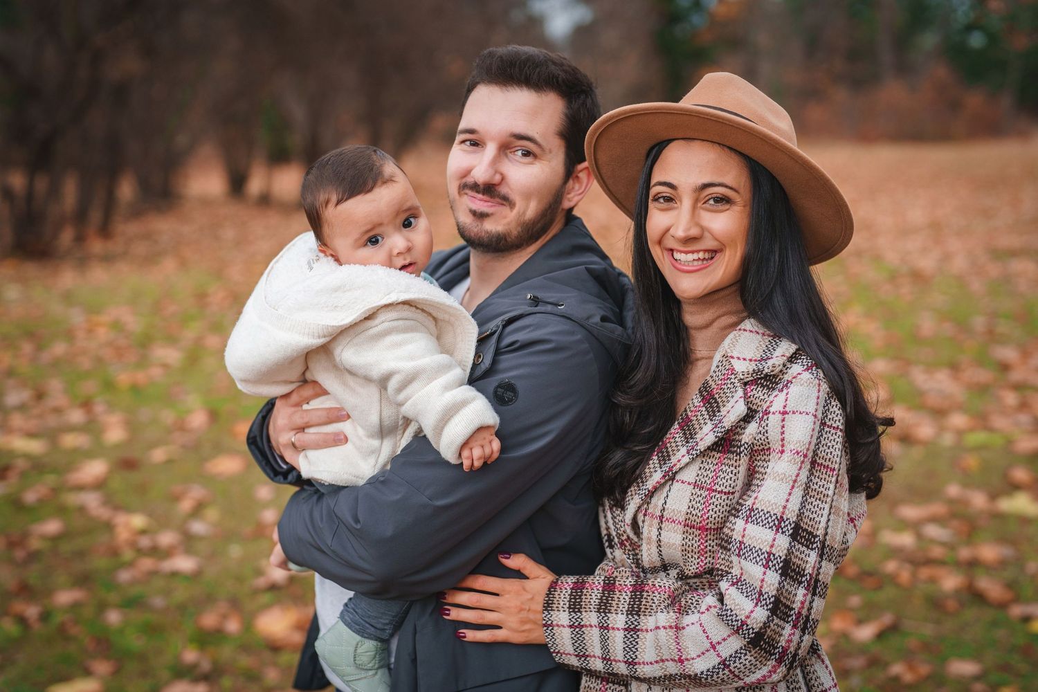 Family poses together outdoors on fallen autumn leaves with stylish brown hat and plaid dress.