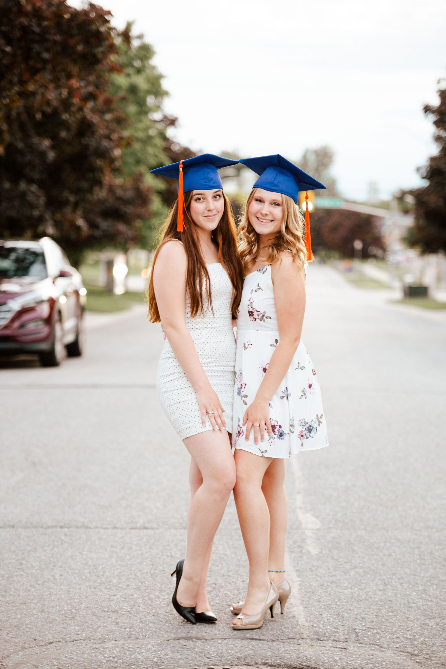 Two young women in white dresses and blue graduation caps pose together on a suburban street.