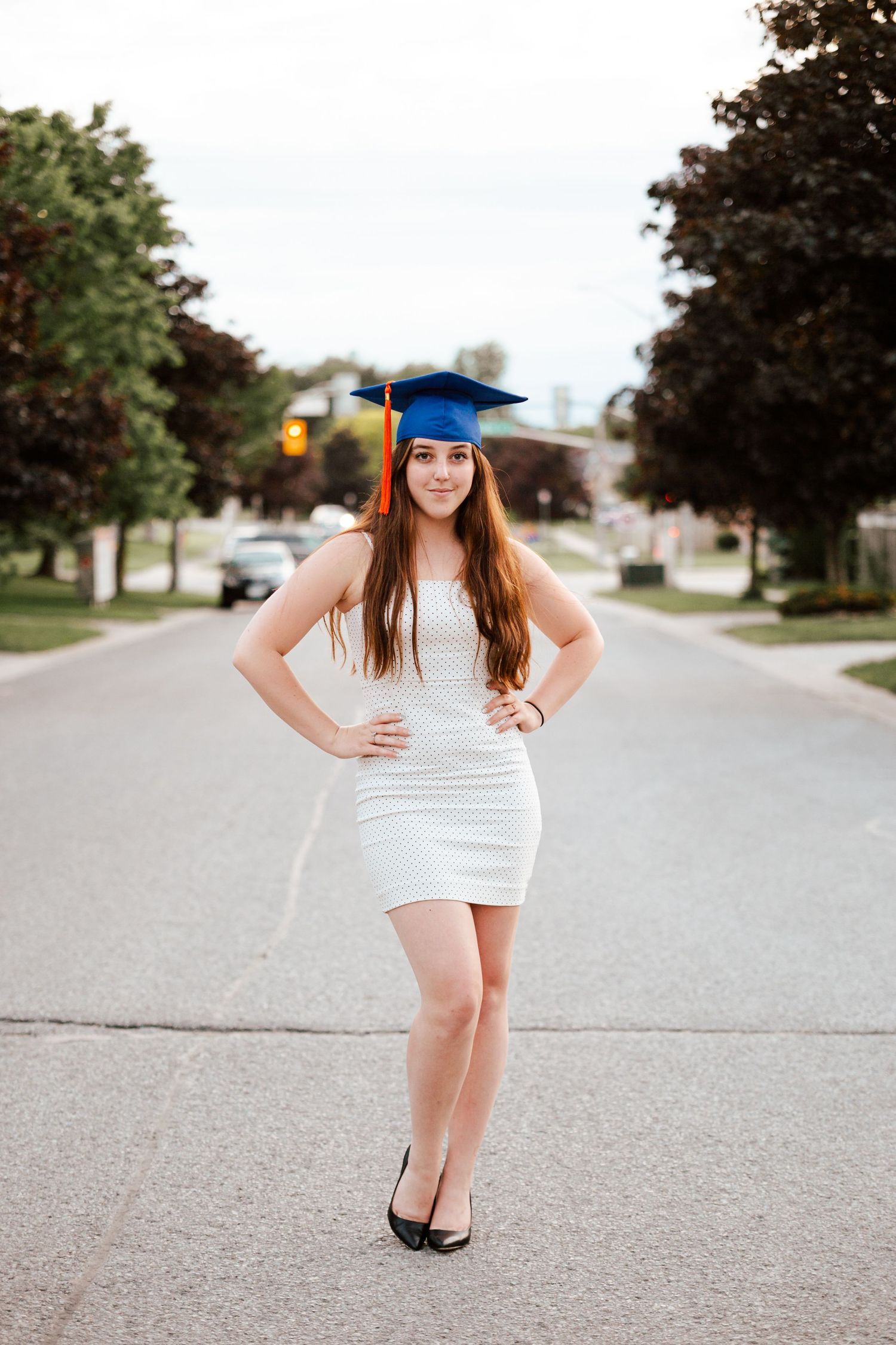 Young woman in white dress and blue graduation cap poses confidently on a tree-lined street.