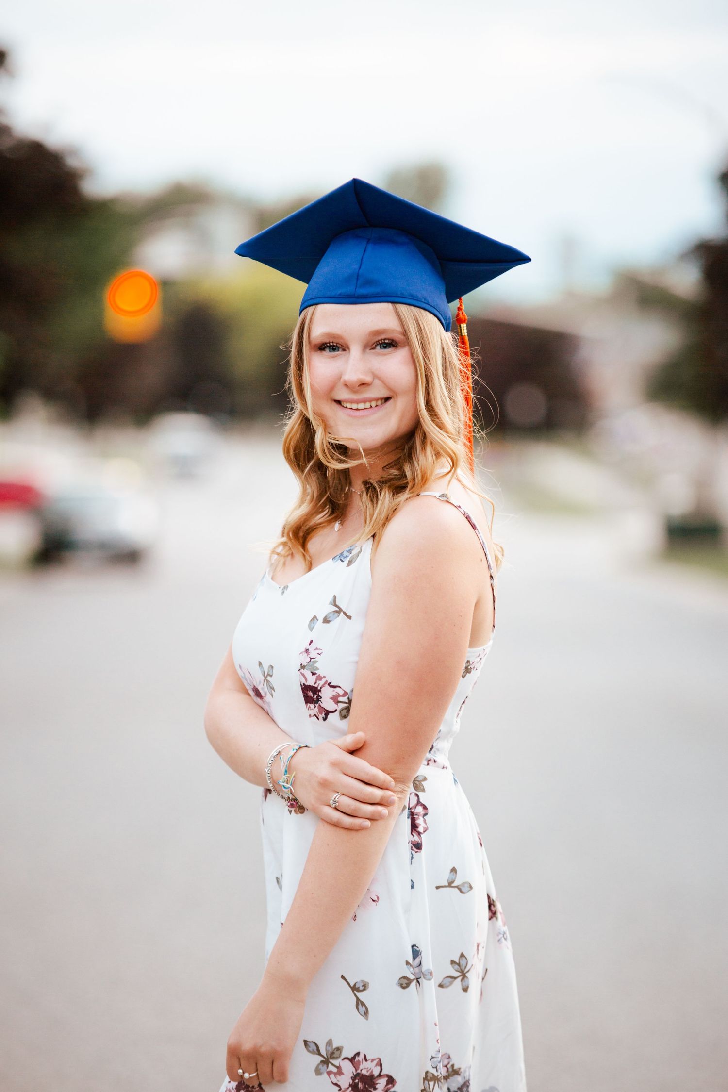 Smiling blonde graduate in floral dress and blue cap poses outdoors on a suburban street.