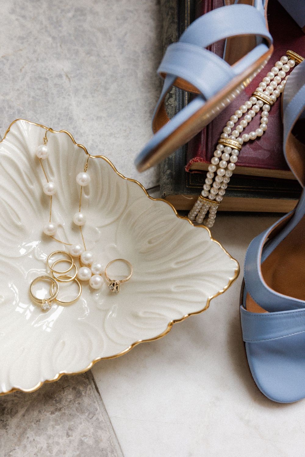 Wedding accessories including blue shoes, pearl jewelry and rings displayed on a decorative white shell dish.