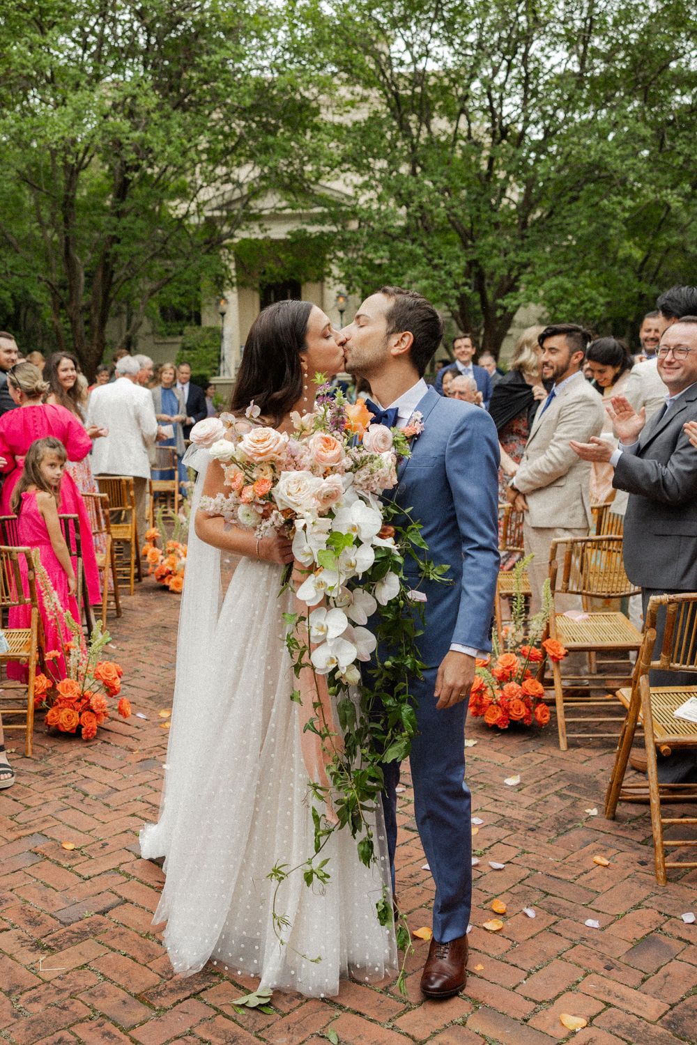 Outdoor wedding ceremony on brick patio with wooden chairs and guests in colorful attire.