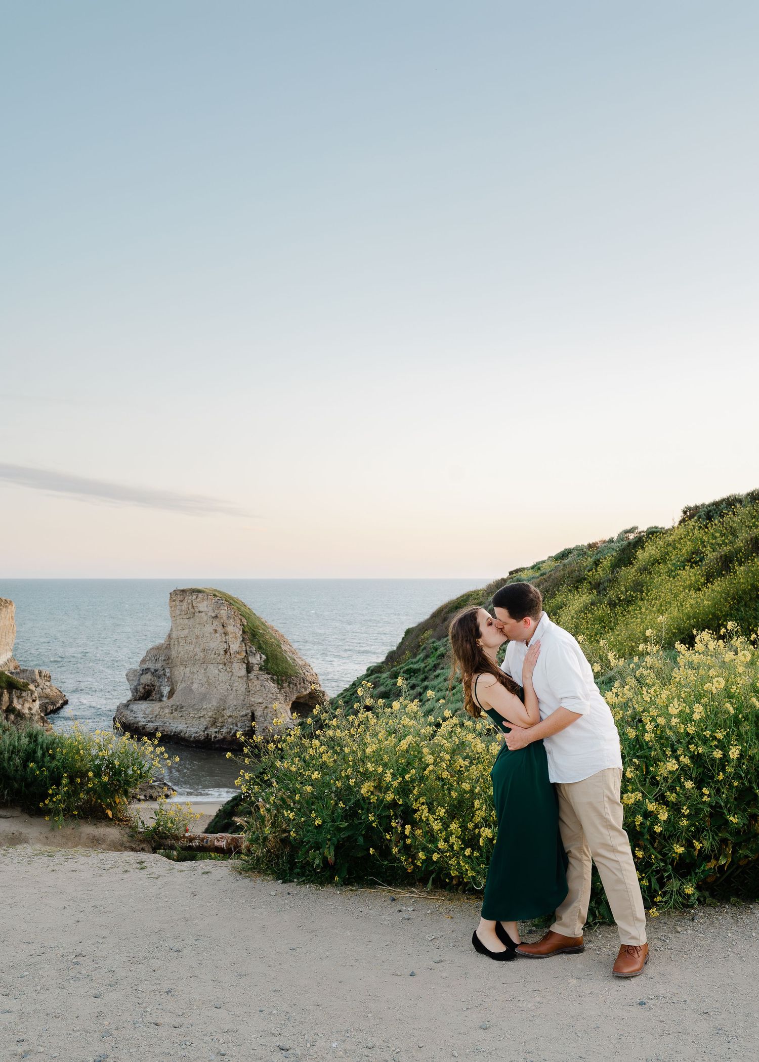 A romantic embrace near coastal cliffs surrounded by yellow wildflowers at sunset.