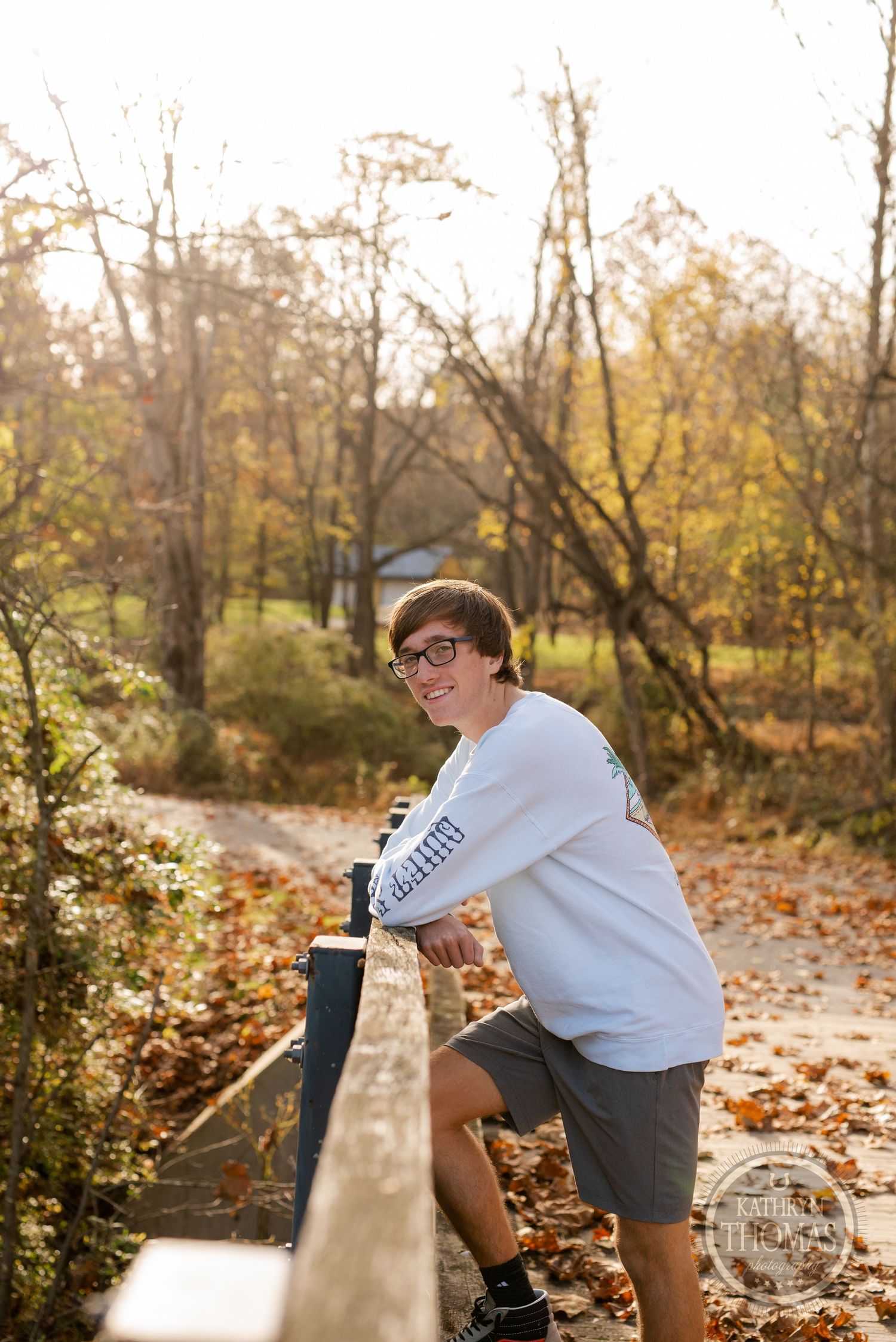 Person leaning on wooden railing of bridge surrounded by fall foliage and golden sunlight.