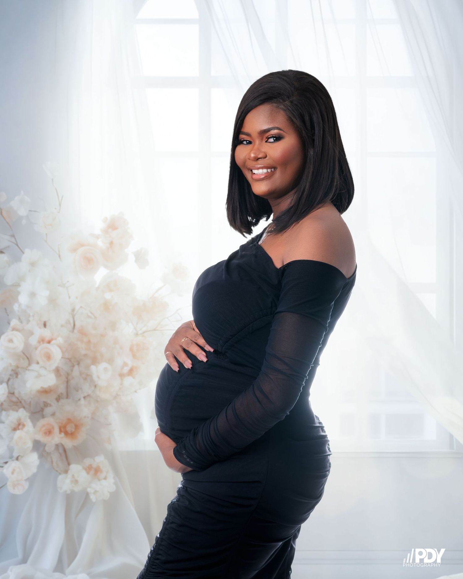 A maternity photo featuring an elegant black off-shoulder dress against a white floral backdrop.