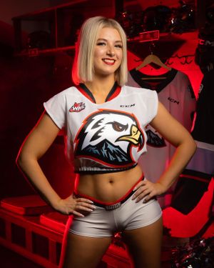 Ice hockey professional dancer in white crop top uniform with Portland Winterhawks logo poses against red backdrop.