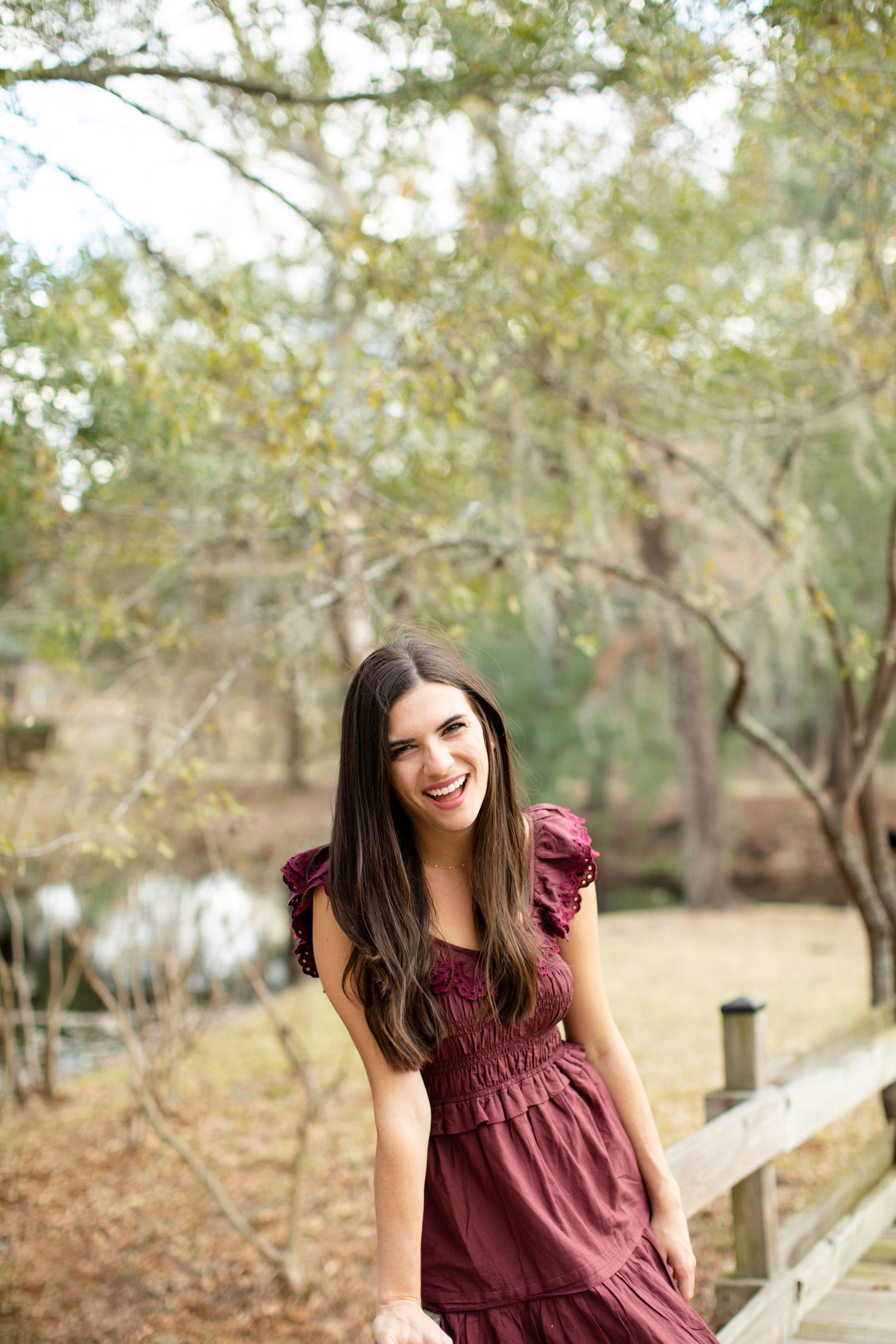 Woman in maroon dress poses happily near a wooden fence in a natural setting.