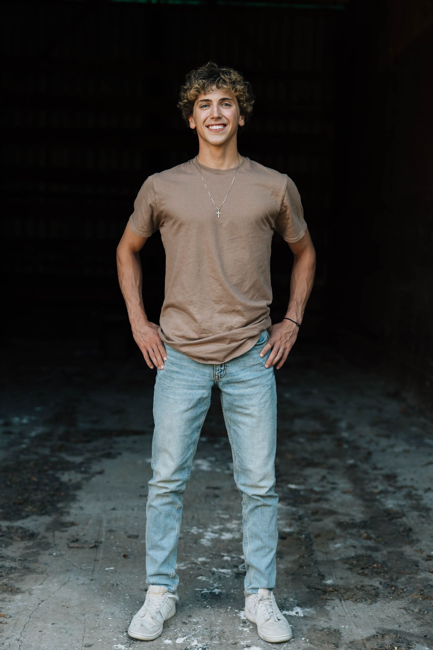 Young man in brown t-shirt and jeans stands confidently in dark background.