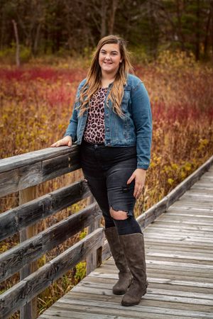 Woman in denim jacket and boots stands casually on a wooden boardwalk surrounded by fall foliage.