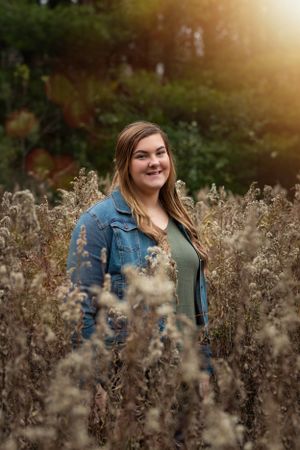 A figure stands among tall dried wildflowers at sunset wearing a denim jacket in a natural outdoor setting.