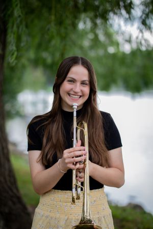 A musician poses with a trumpet outdoors under willow trees by a lake wearing black and yellow.