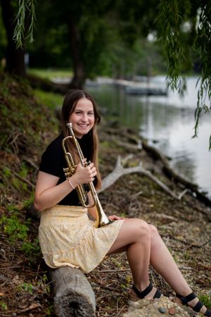 Musician with a trumpet poses near a waterfront under weeping willow branches in casual dress and boots.