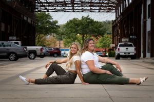 Friends sitting back-to-back on pavement under covered parking structure.