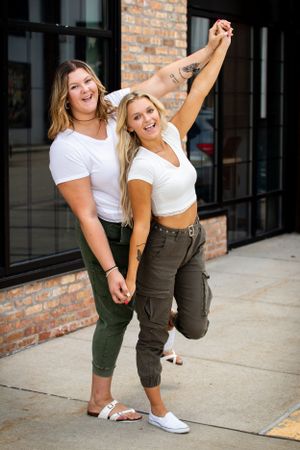 Two friends in matching white tops and cargo pants pose playfully on sidewalk.