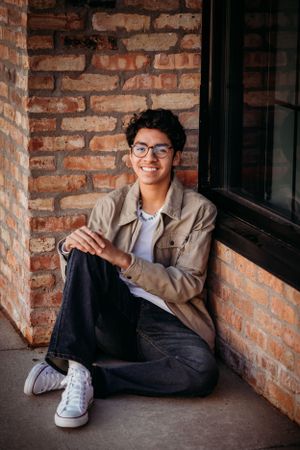 Relaxed portrait sitting against brick wall wearing casual jacket and glasses.