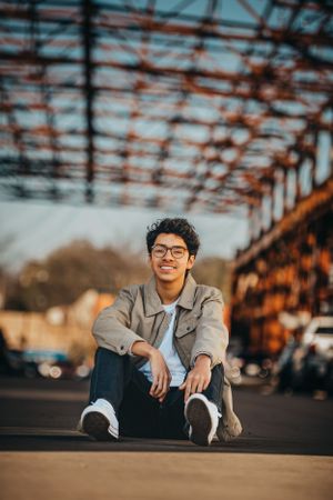 Young person in casual attire sitting on ground near industrial bridge structure at sunset.