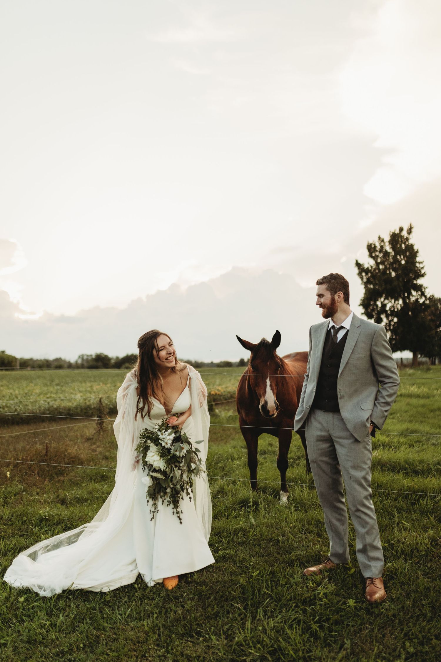 A wedding couple poses in a field with a horse against a sunset sky.