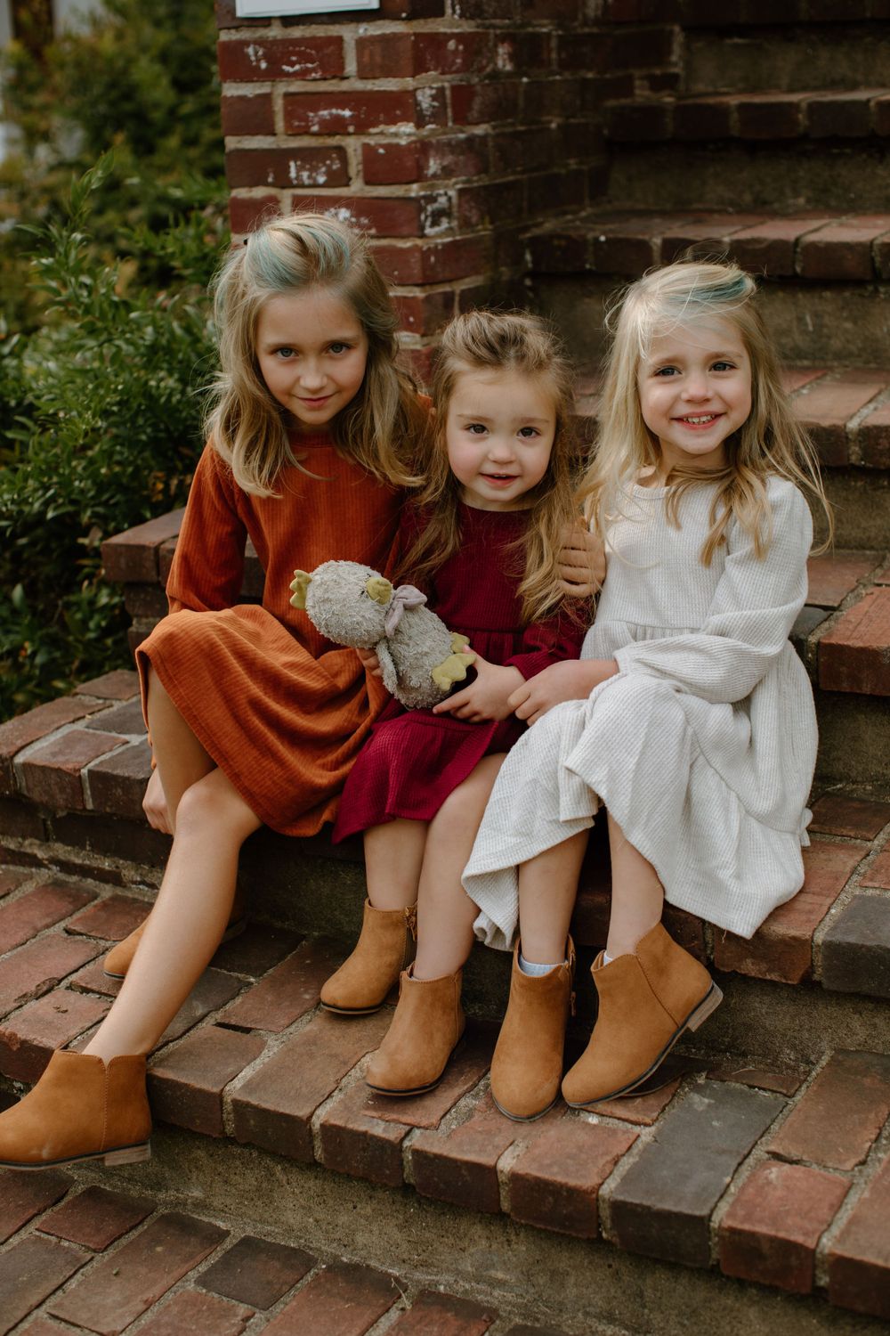 Three young children sitting together on brick steps wearing fall-colored clothing and boots.