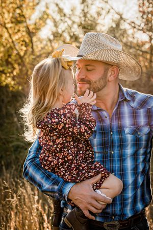 A cowboy in a plaid shirt embraces a small child in a floral dress during a golden sunset.
