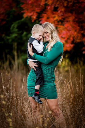 Woman in green dress holds young boy against vibrant fall foliage background.