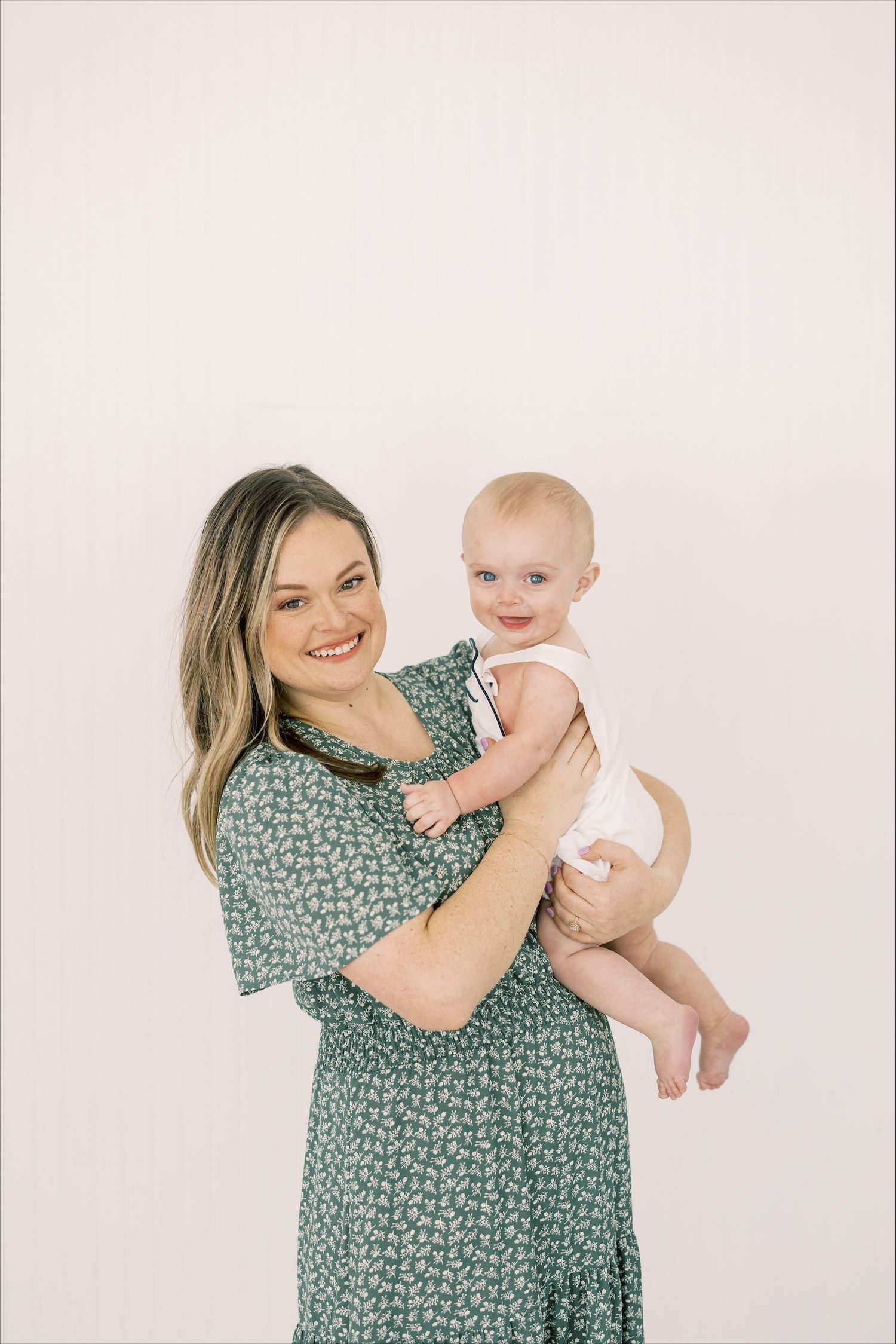 A woman in a green floral dress holds and plays with a happy baby against a light pink backdrop.