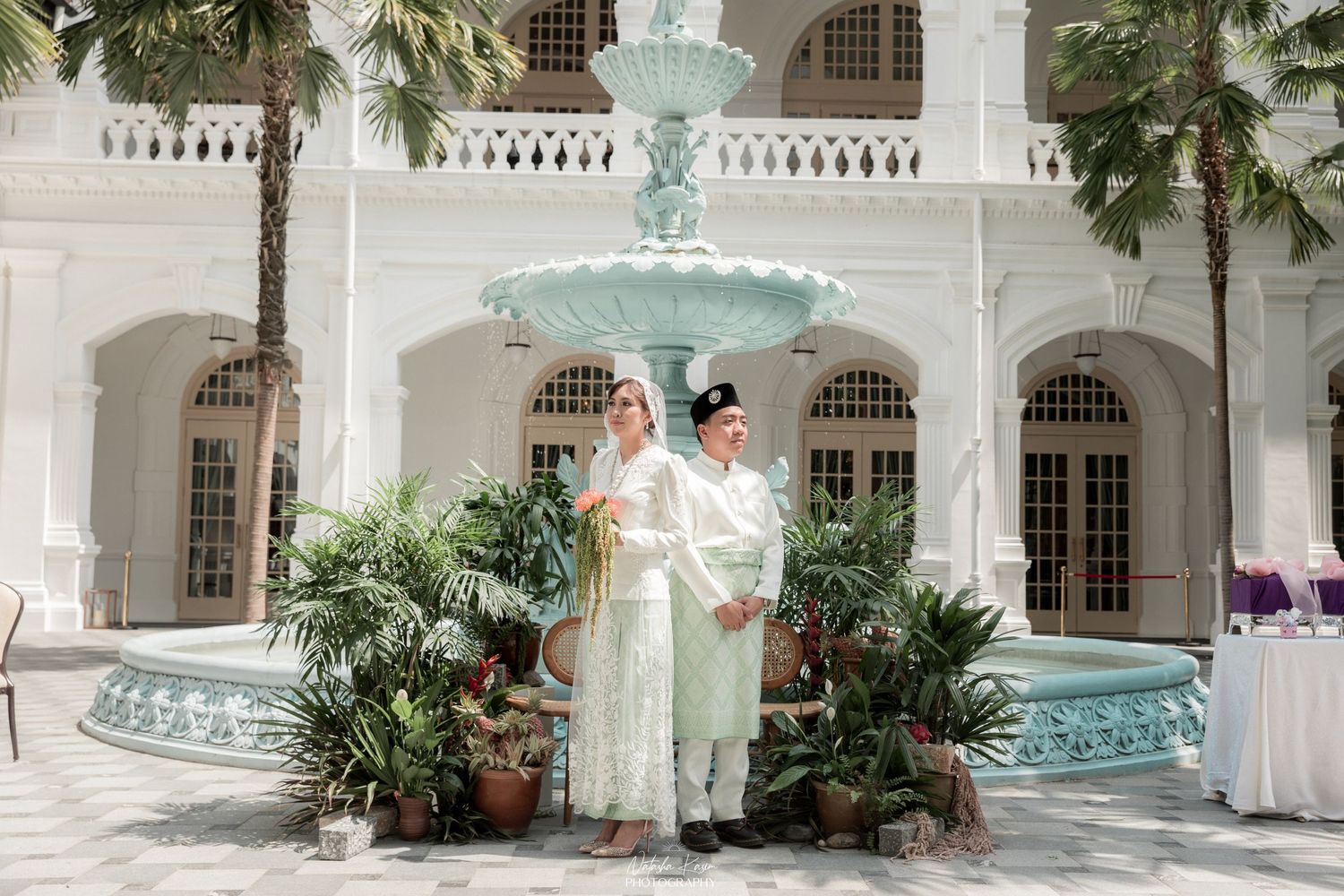 A couple poses in front of an elegant mint-colored fountain surrounded by tropical plants and white colonial architecture.