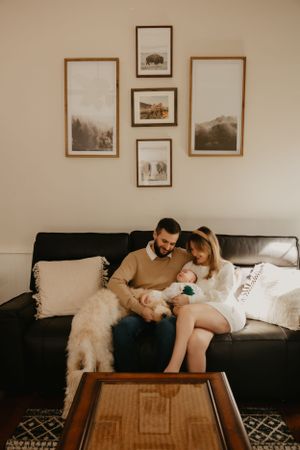 Family relaxes on couch with baby and dog beneath framed wall art.