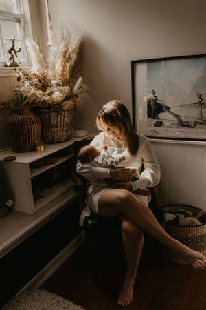 Mother holds newborn baby in cozy room with dried flower decorations.