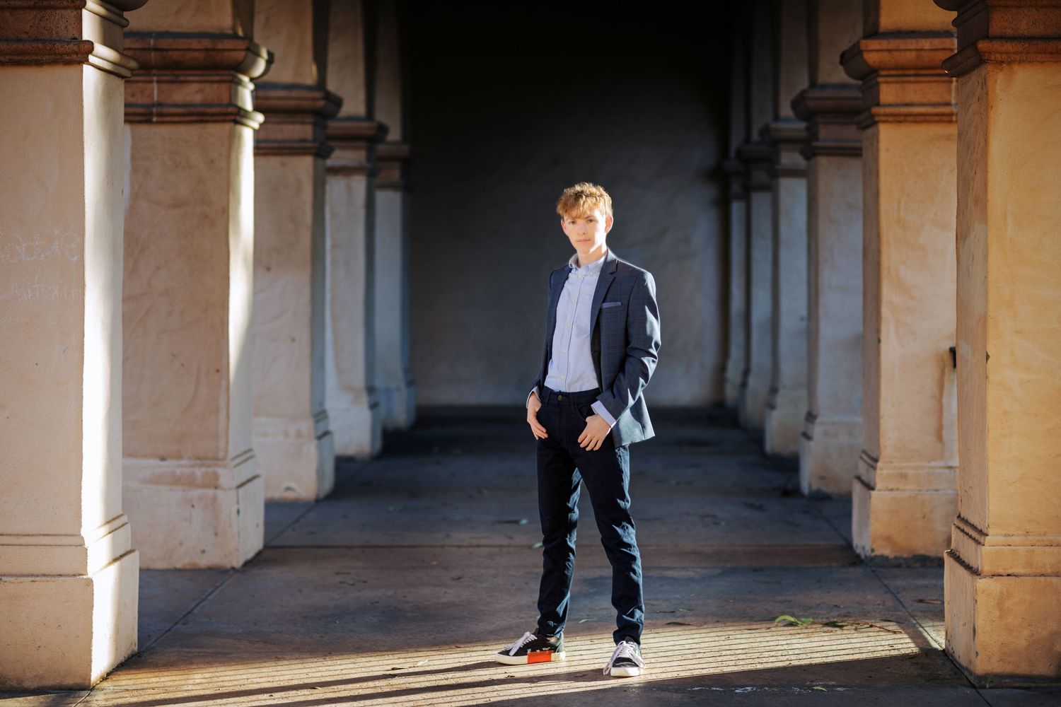 A high school senior standing in shadowy colonnade wearing casual blazer and jeans.
