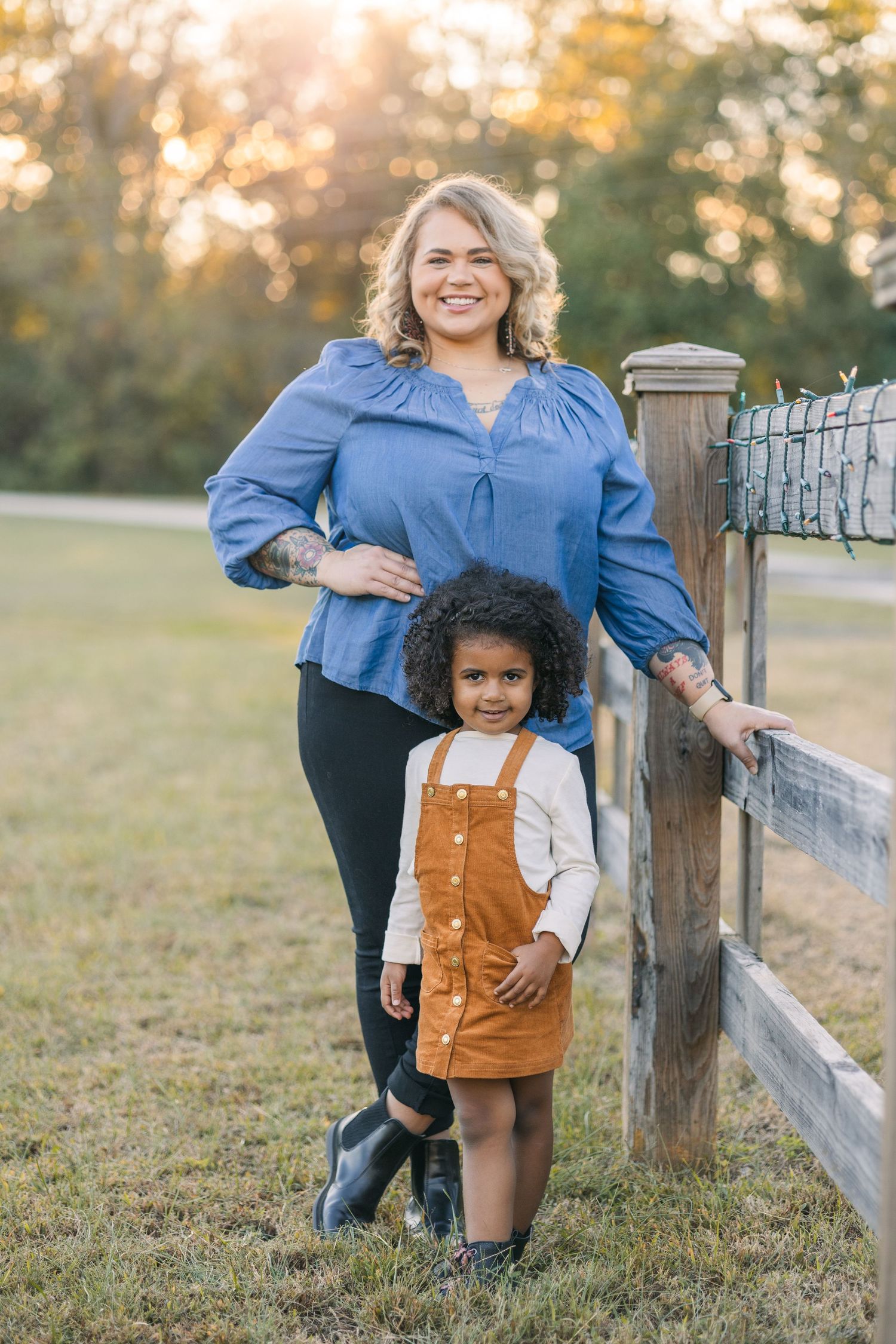mother in a blue shirt stands behind daughter in field in an orange outfit during golden hour