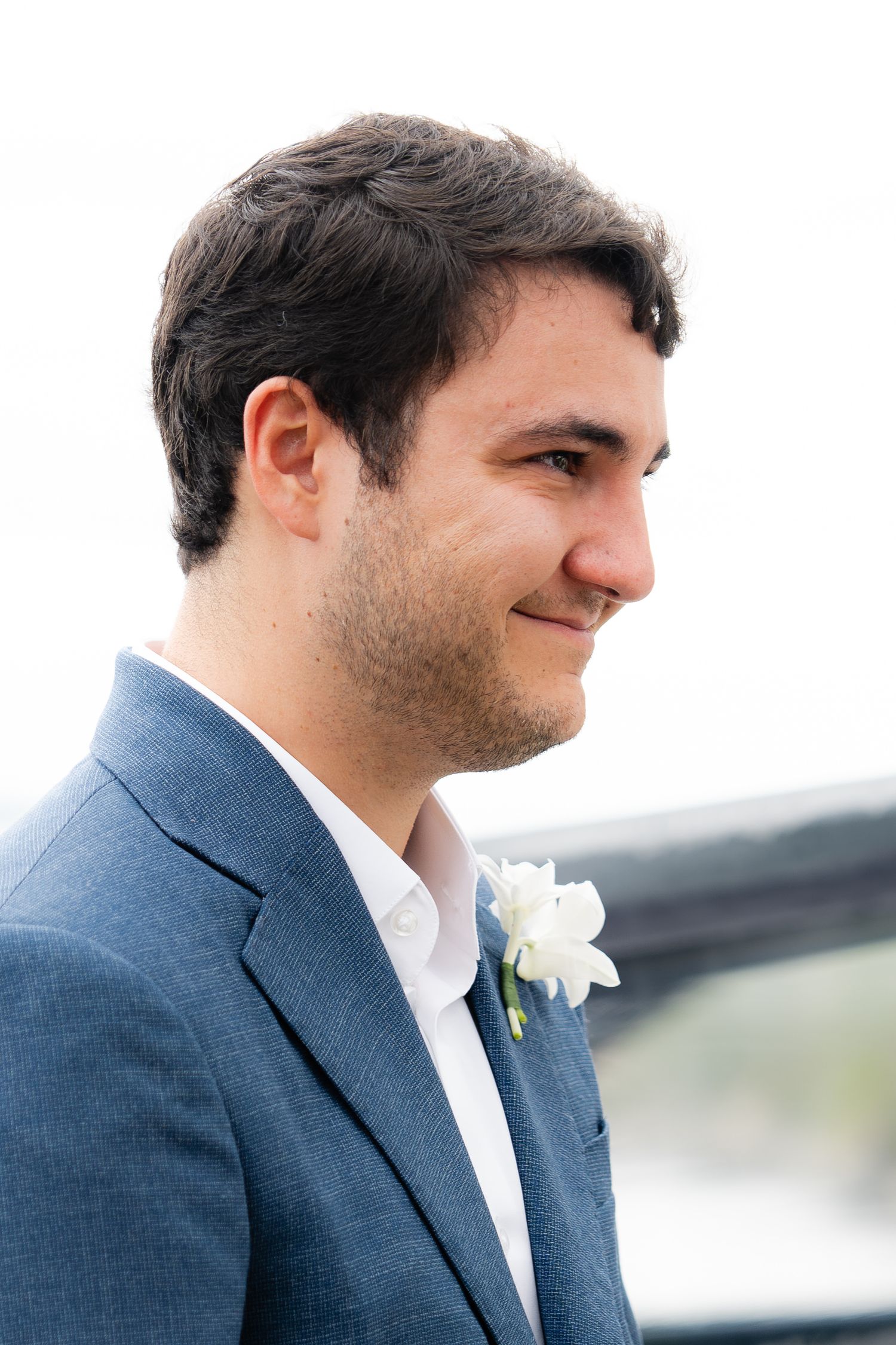 Groom in navy suit with white boutonniere smiles during outdoor wedding.