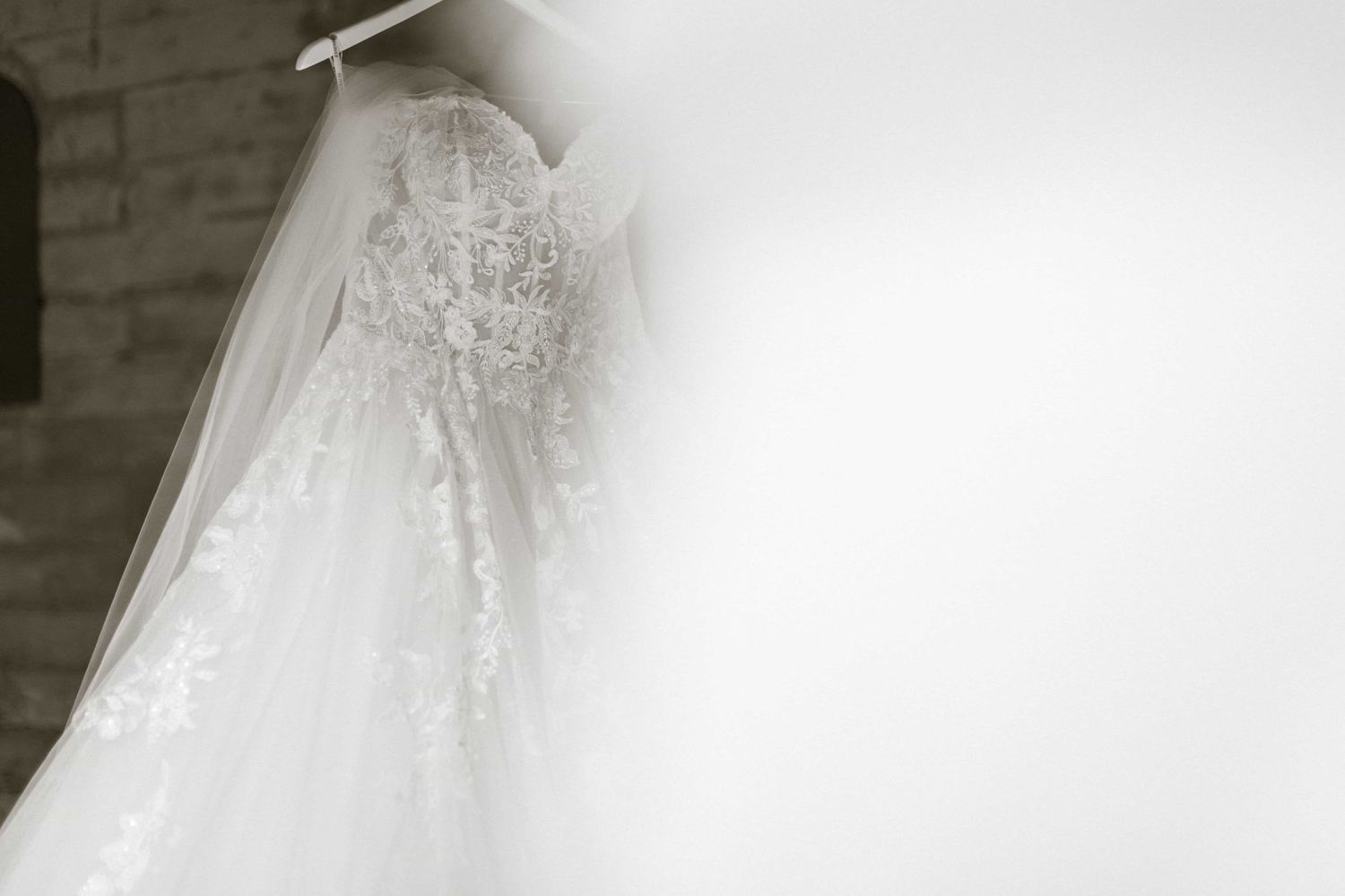 Elegant white lace wedding gown with intricate floral embroidery hanging against a rustic brick wall.