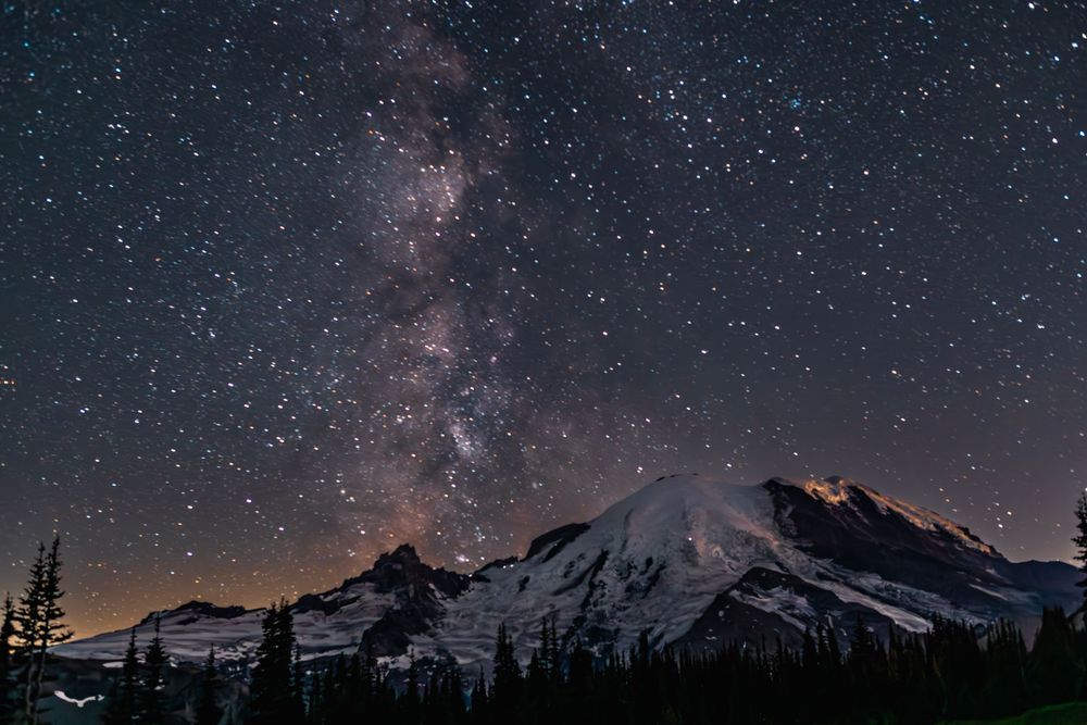 Stunning night sky with Milky Way galaxy arching over snow-capped mountain peaks and silhouetted evergreen trees.