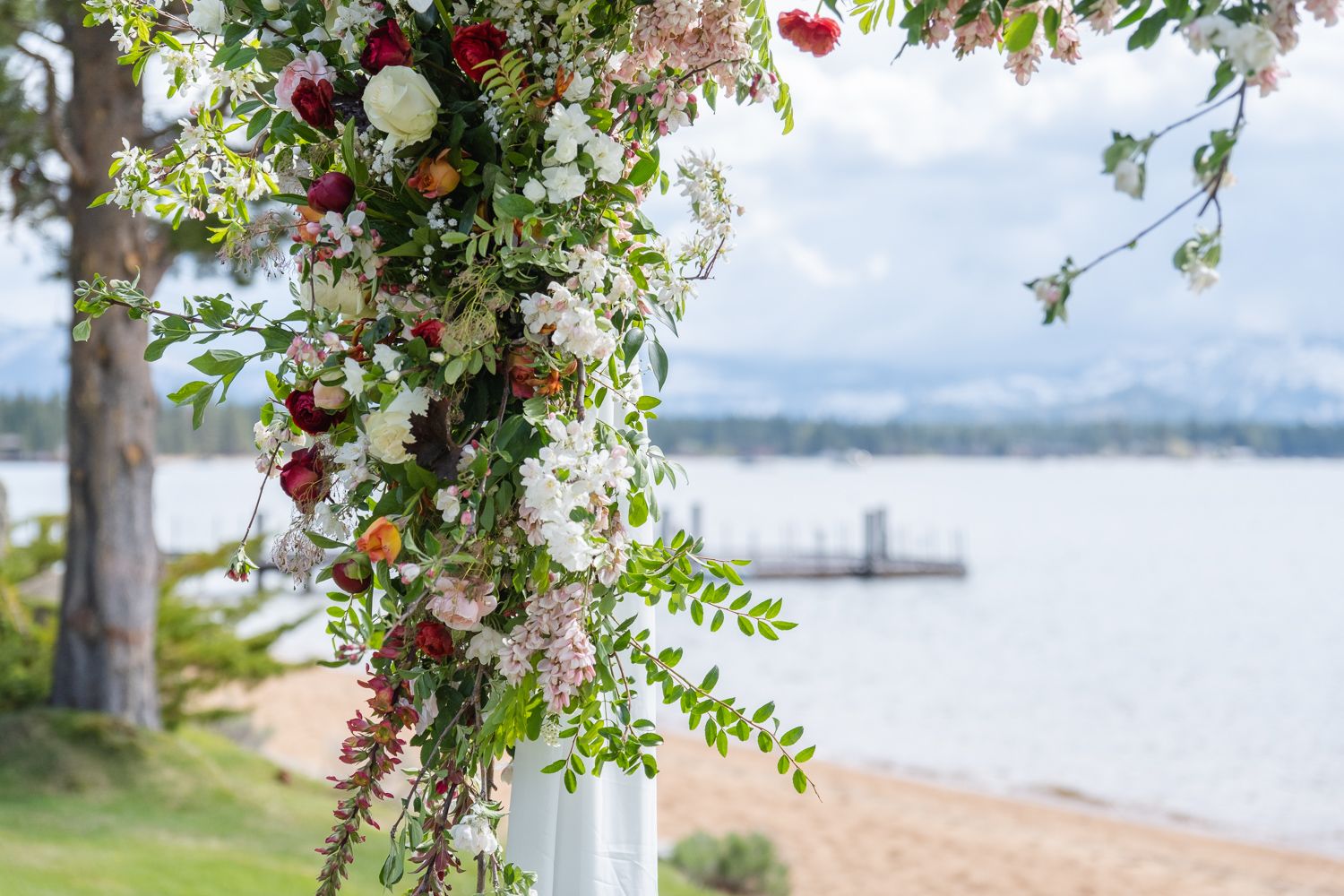 An elegant floral arch with red and white flowers decorates a waterfront ceremony space overlooking a peaceful lake.