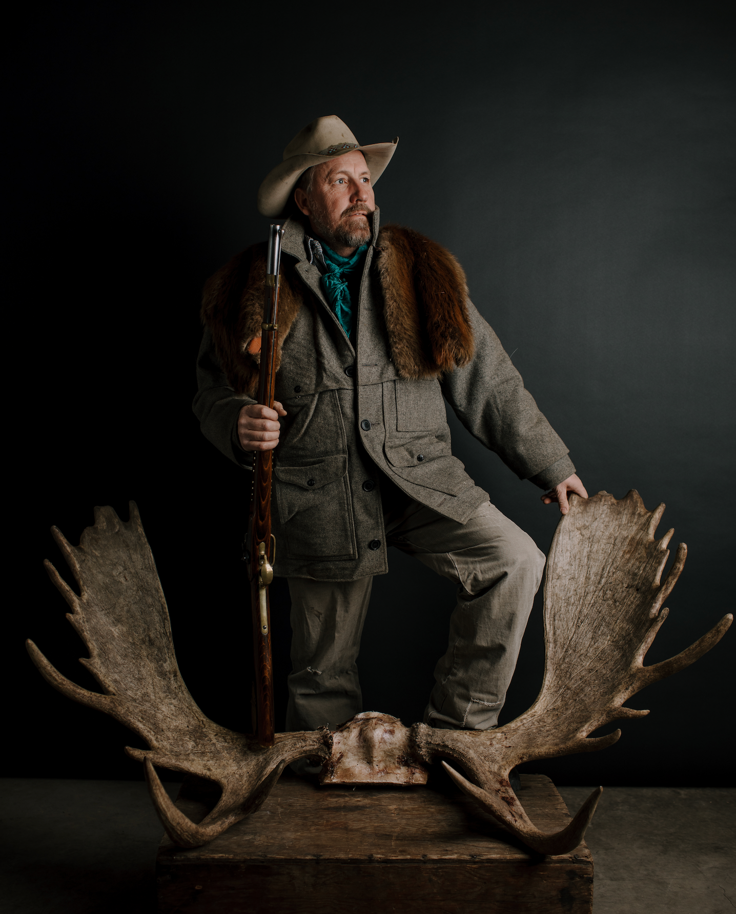 A hunter in outdoor gear stands atop large moose antlers against a dark background.